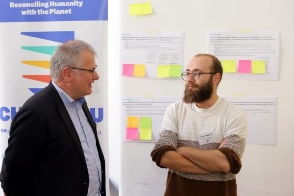 Two participants at the libraries meet-up discussing with the CHARM-EU rollup on the back and a panel with notes and post-its.