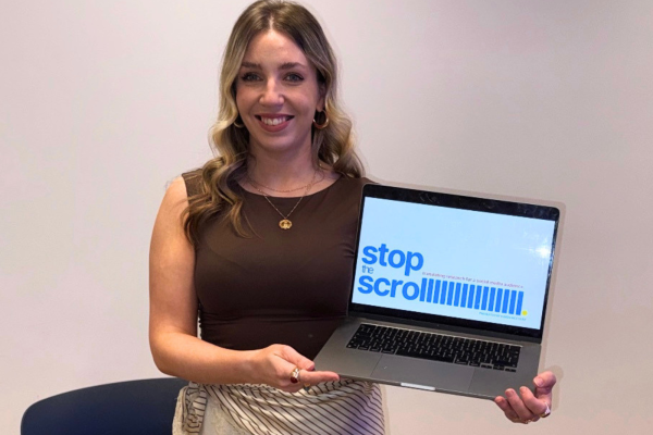 A smiling woman with wavy blonde hair stands against a plain light wall, holding an open laptop towards the camera. She is wearing a brown sleeveless top, necklaces, and a striped wrap around her waist. The laptop screen displays a graphic with the bold blue text "stop the scroll", followed by a series of vertical blue lines and smaller, lighter text that reads "translating research for a social media audience."