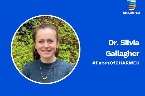 A professional graphic on a solid blue background featuring a circular headshot of Dr. Silvia Gallagher on the left. She is smiling, with dark hair tied back, wearing a blue sweater and a gold necklace, positioned in front of green foliage. To the right of the photo, the white text reads "Dr. Silvia Gallagher" followed by the hashtag "#FacesOfCHARMEU." The top right corner contains the CHARM-EU logo, which consists of a stylized shield with colorful horizontal stripes.
