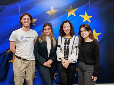 European Commission's Executive Vice-President Roxana Mînzatu with the 3 students and alumni from European Universities alliances with the flag of the European Union on the background.