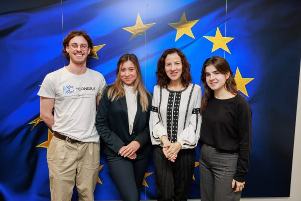 European Commission's Executive Vice-President Roxana Mînzatu with the 3 students and alumni from European Universities alliances with the flag of the European Union on the background.