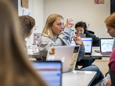 Students with their laptops. One of them is explaining somthing with her hands to her colleagues. Other studetns are looking at their computers.