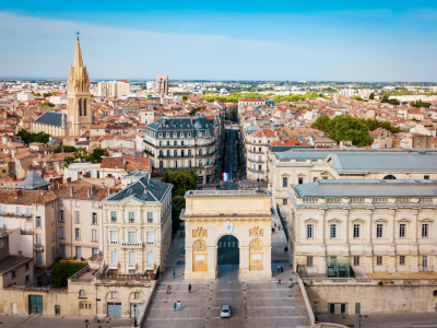 An aerial, high-angle view of the Arc de Triomphe in Montpellier, France (Porte du Peyrou), a grand stone triumphal arch situated at the end of a wide pedestrian promenade. A single white car drives through the arch, and a few pedestrians are scattered across the paved plaza. The arch aligns with a long street that cuts through the city, where a French flag flies in the distance. The surrounding cityscape features historic European buildings with terracotta tiled roofs, stone facades, and a prominent church spire rising on the left. The sky above is clear and bright blue.