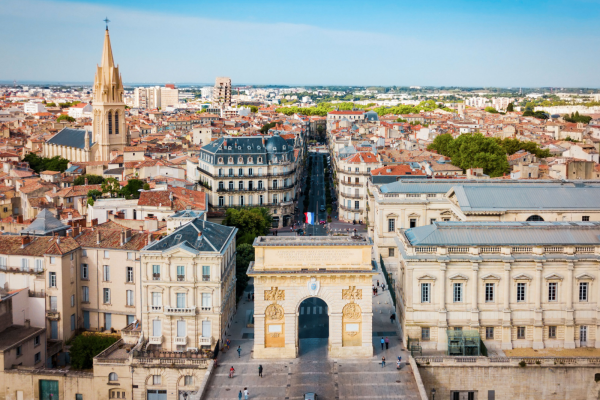 An aerial, high-angle view of the Arc de Triomphe in Montpellier, France (Porte du Peyrou), a grand stone triumphal arch situated at the end of a wide pedestrian promenade. A single white car drives through the arch, and a few pedestrians are scattered across the paved plaza. The arch aligns with a long street that cuts through the city, where a French flag flies in the distance. The surrounding cityscape features historic European buildings with terracotta tiled roofs, stone facades, and a prominent church spire rising on the left. The sky above is clear and bright blue.