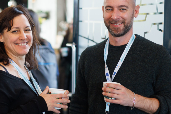 Here is an alternative text description for the image "Peer Coaching - Thrive Together.jpg": **Alt Text:** A man and a woman stand close together smiling at the camera in an indoor setting. Both are wearing lanyards and holding white mugs. The woman on the left has dark brown hair and is wearing a black shirt, while the bald man on the right has a short beard and is wearing a dark grey sweater. The background is slightly blurred with other people visible.
