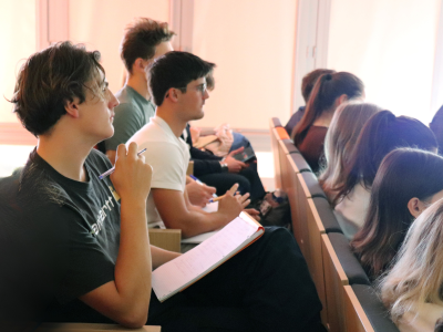 A row of students seated in tiered wooden desks, taking notes in an auditorium or classroom. In the foreground, a young man with curly brown hair wearing a black shirt and glasses holds a pen and an open notebook, looking towards the left, outside the frame. Further back, another student in a white shirt is also looking attentively, while a third is looking down at a mobile device. The background is brightly lit by a window, creating a lens flare and obscuring the faces of others. The focus is primarily on the foreground student and his open notebook.