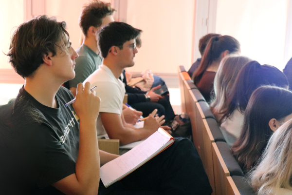 A row of students seated in tiered wooden desks, taking notes in an auditorium or classroom. In the foreground, a young man with curly brown hair wearing a black shirt and glasses holds a pen and an open notebook, looking towards the left, outside the frame. Further back, another student in a white shirt is also looking attentively, while a third is looking down at a mobile device. The background is brightly lit by a window, creating a lens flare and obscuring the faces of others. The focus is primarily on the foreground student and his open notebook.
