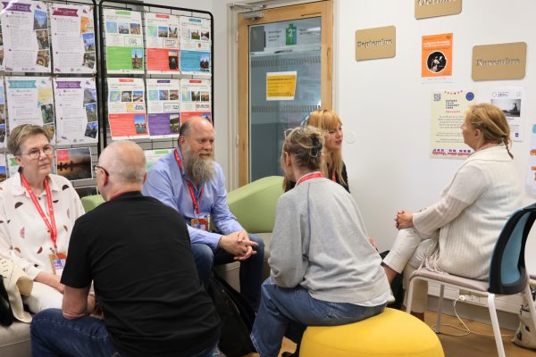 A group of six people sit in a circle in an office or classroom setting, engaged in conversation. On the left, an older woman with glasses and a patterned blouse sits opposite a man with a long gray beard and a blue button-down shirt. Two individuals sit with their backs to the camera in the foreground—one on a blue pouf and another on a yellow pouf. On the right, two blonde women are seated, one in a white cardigan facing the group. The background features a glass-paned door, a wall with signs for "Septembre," "Octobre," and "Novembre," and a display rack filled with colorful informational brochures.