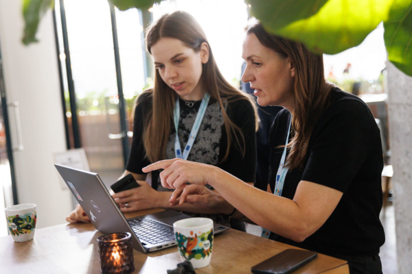 Two colleagues sitting at a wooden table in a bright, modern indoor space work together on a laptop. The person on the right, wearing a black shirt, points at the laptop screen while explaining something. The person on the left, holding a smartphone, looks closely at the screen. Both are wearing light blue event lanyards. On the table are two patterned coffee mugs, a smartphone, and a small lit candle holder, with green leaves softly blurring the top right foreground.