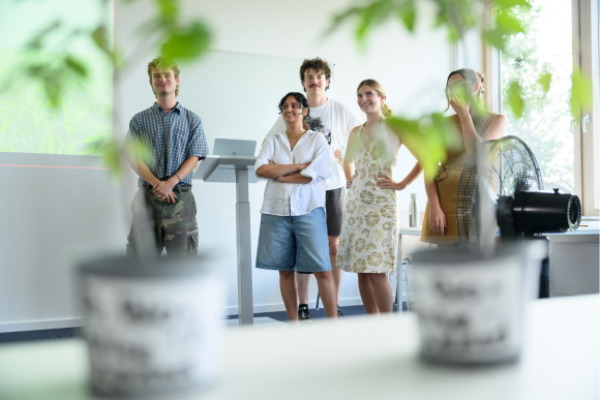 Group of students standing in a classroom during a sustainability-related presentation, viewed through foreground plants.