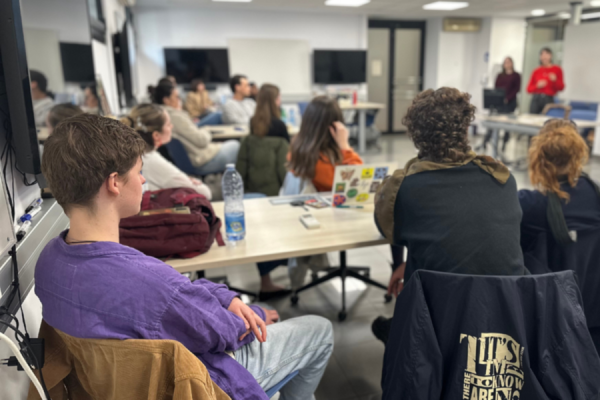 Participants seated around tables attending a workshop session, listening to presenters speaking at the front of a meeting room.